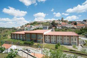 Complexo moderno com cobertura vermelha integrado em paisagem rural com aldeia ao fundo e céu azul.