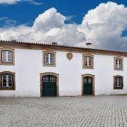 Edifício de fachada branca com detalhes em pedra, portas e janelas em verde, sob céu azul.