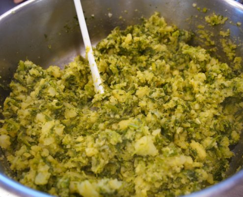 Mistura de ingredientes verde-amarelados em panela de metal com colher de madeira