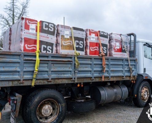 Camião com caixa de carga azul transportando caixas de material de construção com marcas CS, fixas com cintas amarelas e laranja.